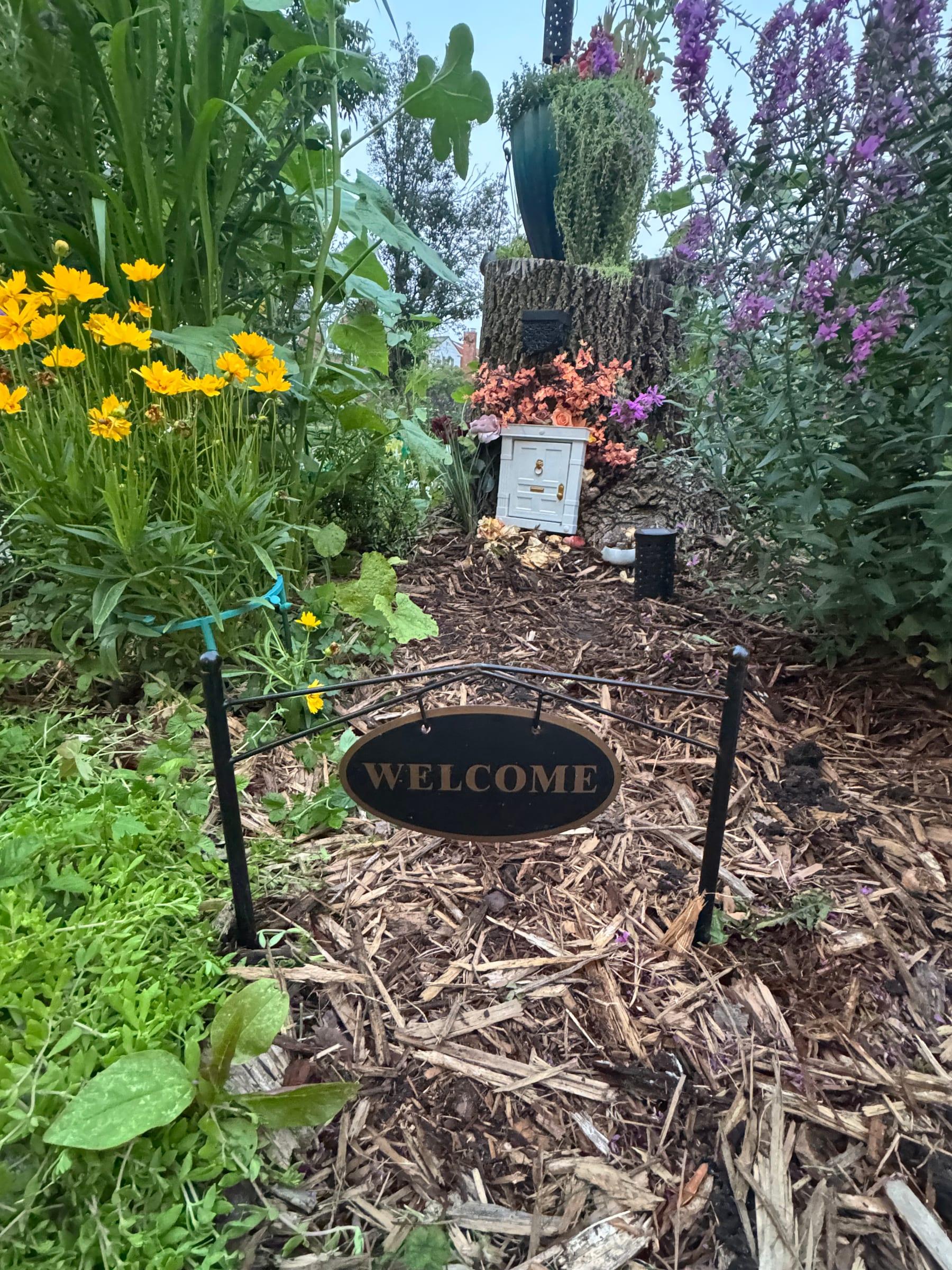 A mulched garden path lined with yellow flowers leading to a welcome sign